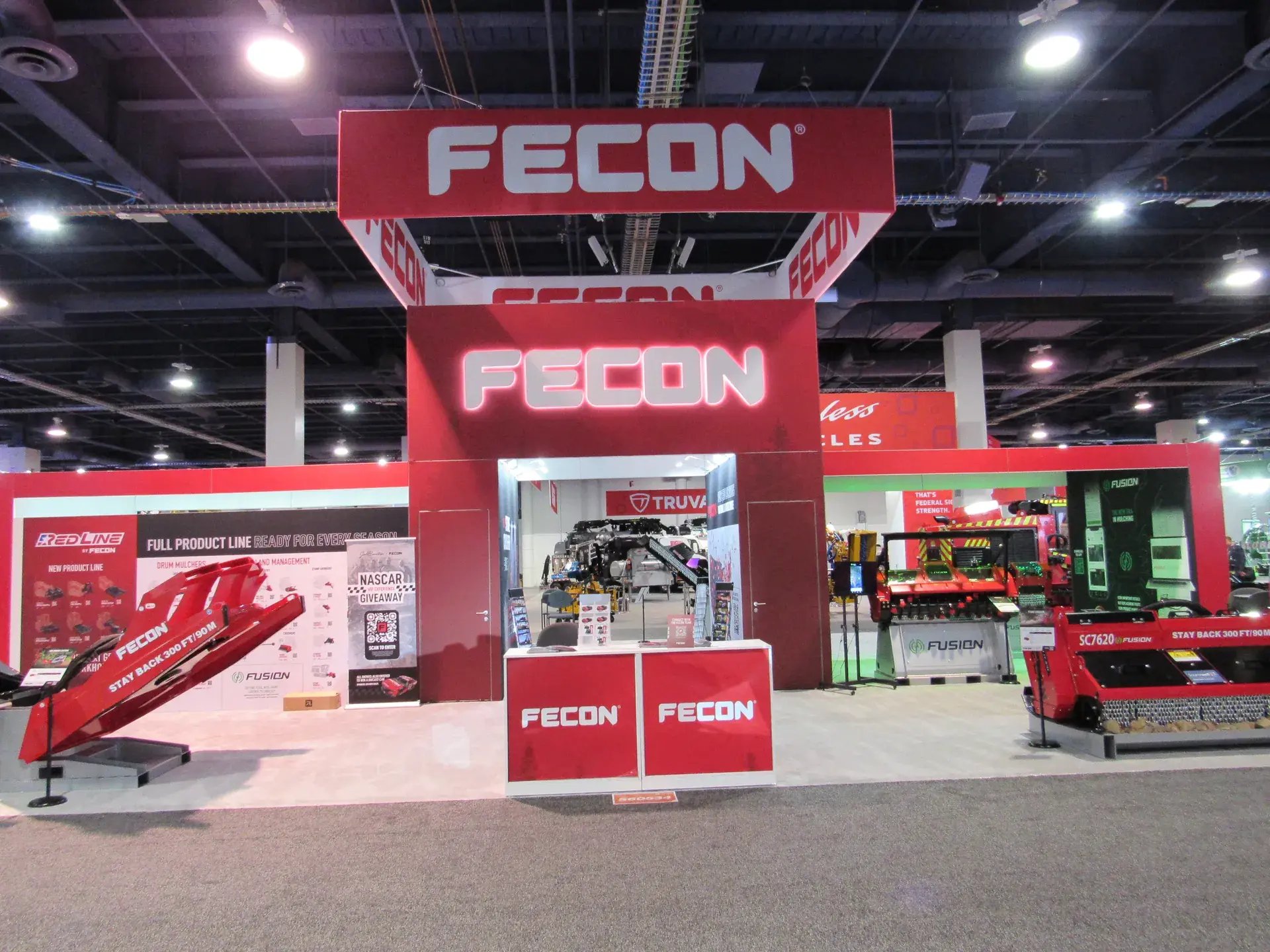 Fecon 40x80 island exhibit at ConExpo with a central tower and 20x20 overhead hanging sign above red forestry equipment